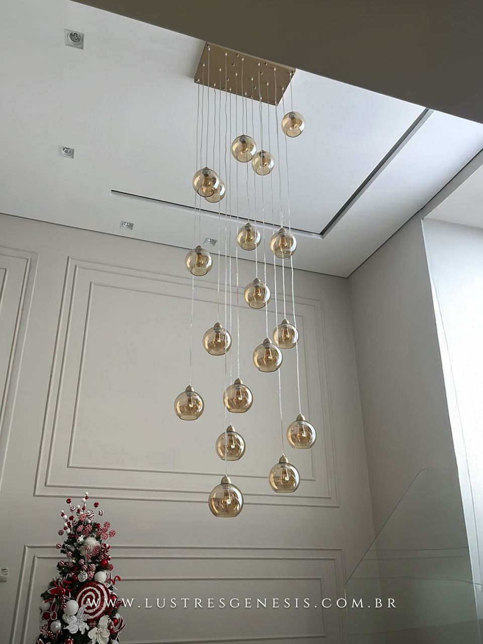 Lustre pendente Sarvah com 21 globos conhaque instalado em ambiente de pé direito duplo, ao lado de uma árvore de Natal decorada e urso de pelúcia.